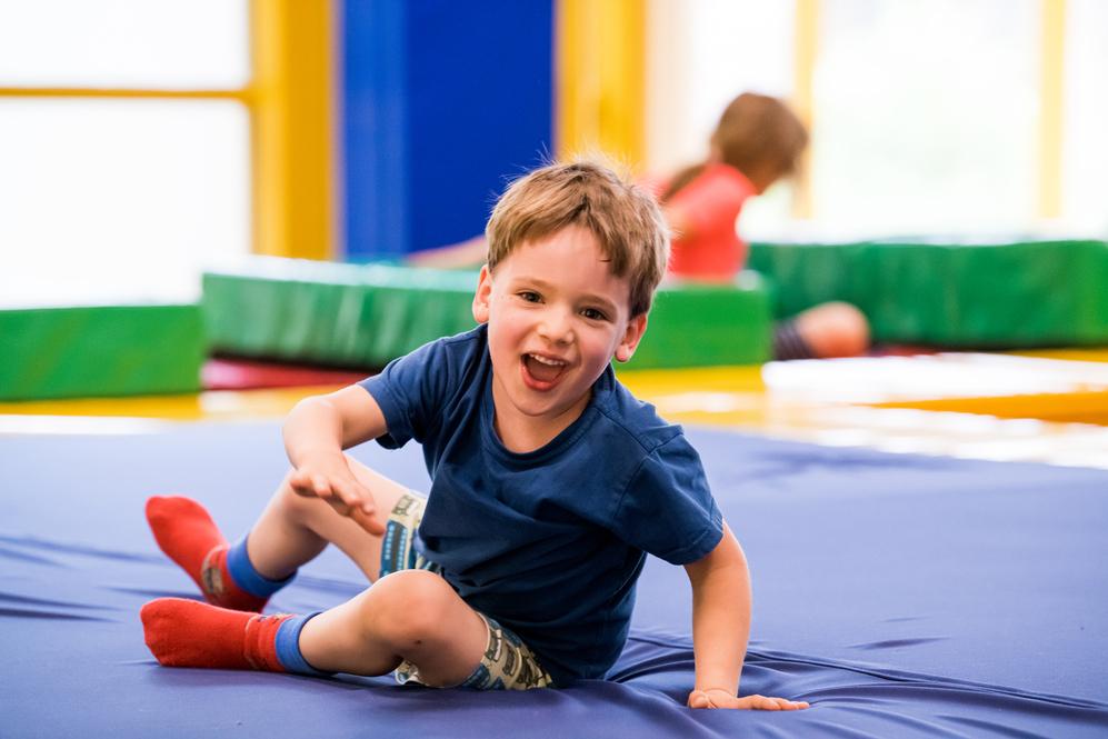 Ein fröhlicher Junge spielt auf einer blauen Matte in einer Freizeitstätte, umgeben von bunten Matten.