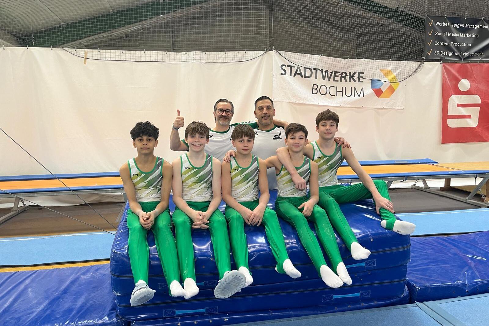 Gruppe von sechs Jungen in grünen Gymnastikanzügen auf einem blauen Kästchen, begleitet von einem Trainer im Hintergrund.
