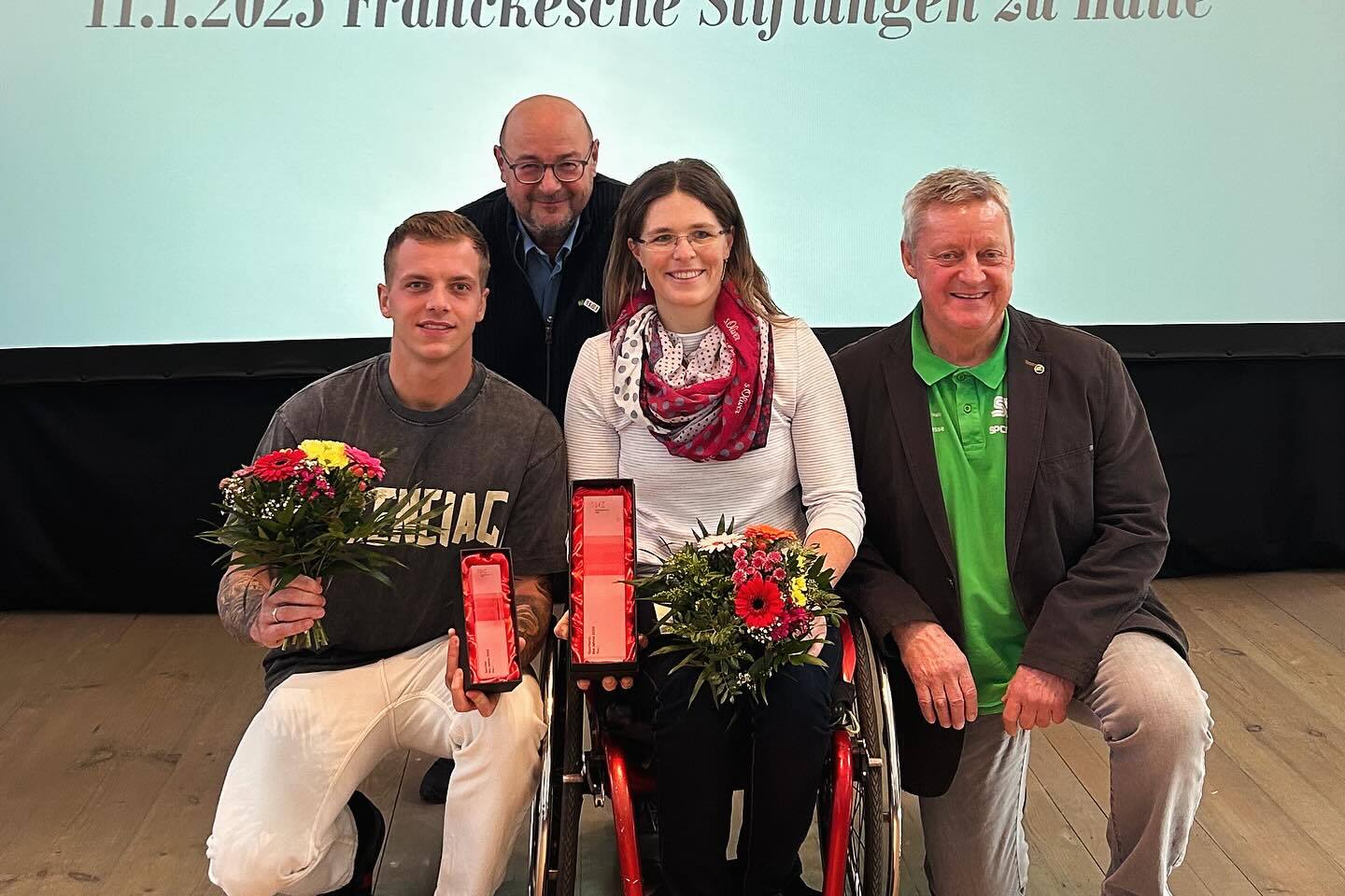 Vier Personen posieren mit Blumensträußen und Auszeichnungen vor einem Präsentationshintergrund bei einer Sportveranstaltung.