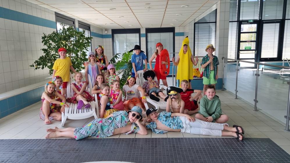 Gruppenfoto von Kindern in bunten Kostümen und Sonnenaccessoires, posierend im Schwimmbad mit Pflanzen im Hintergrund.
