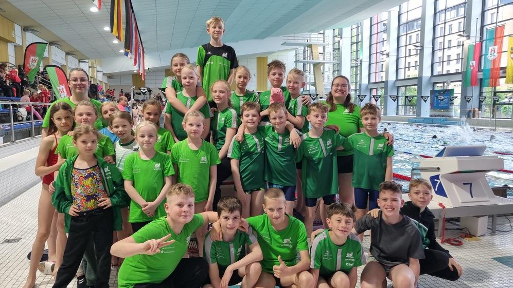 Gruppe junger Schwimmer in grünen T-Shirts posiert fröhlich am Beckenrand eines Schwimmbades.