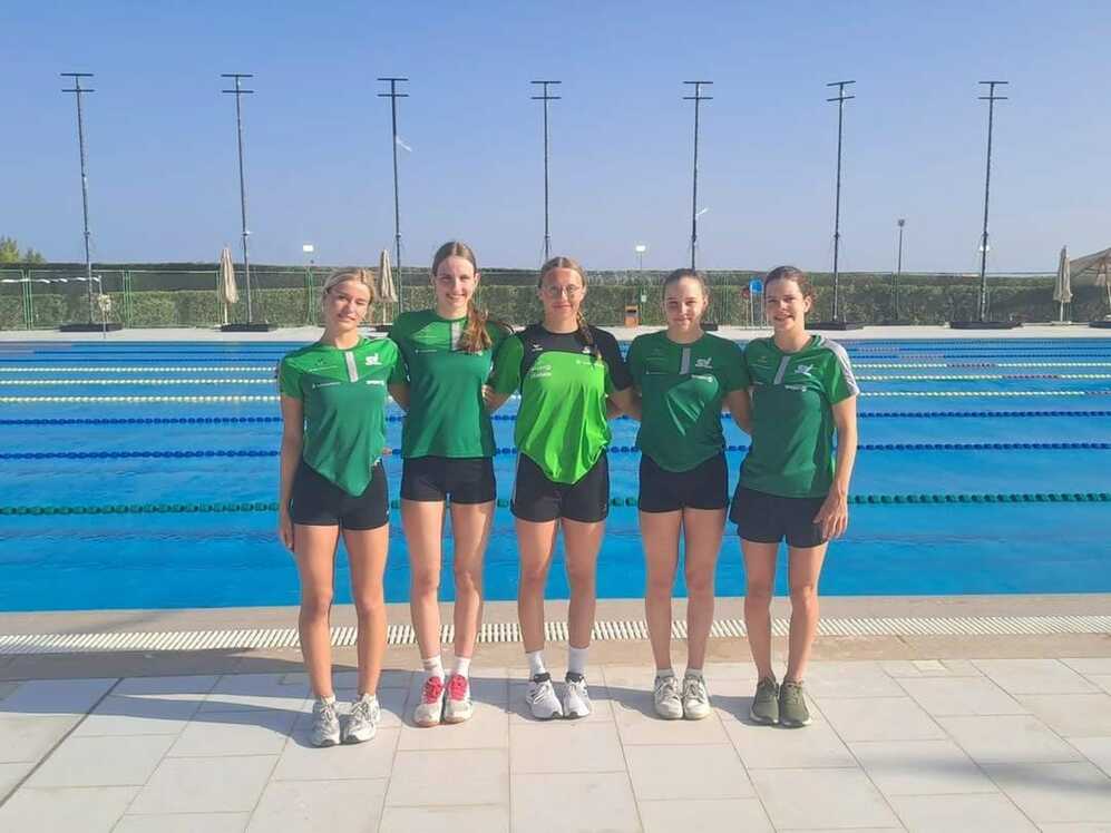 Fünf Schwimmerinnen in grünen T-Shirts stehen am Rand eines Schwimmbeckens unter blauem Himmel.