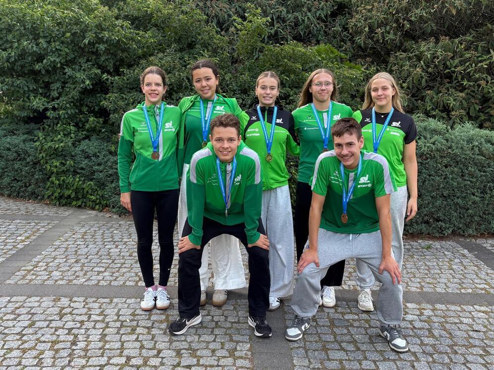 Gruppenfoto von sechs Sportlern in grünen Trikots mit Medaillen, posierend vor einer Hecke auf einem Pflasterweg.