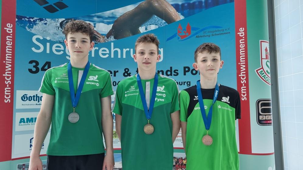 Drei junge Schwimmer mit Medaillen stehen vor einem Banner bei einer Siegerehrung im Schwimmsport.