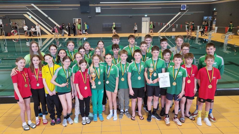Gruppe von Schwimmern in Sportbekleidung mit Medaillen und Pokalen, posierend in einem Schwimmbad.