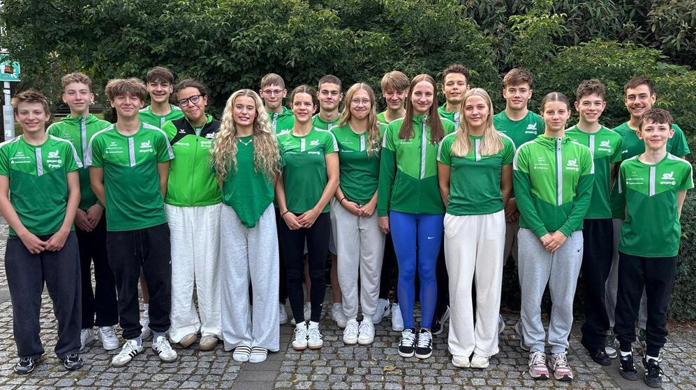 Gruppenbild von 16 jungen Sportlern in grünen Trikots, stehend vor einer grünen Hecke auf einem gepflasterten Weg.