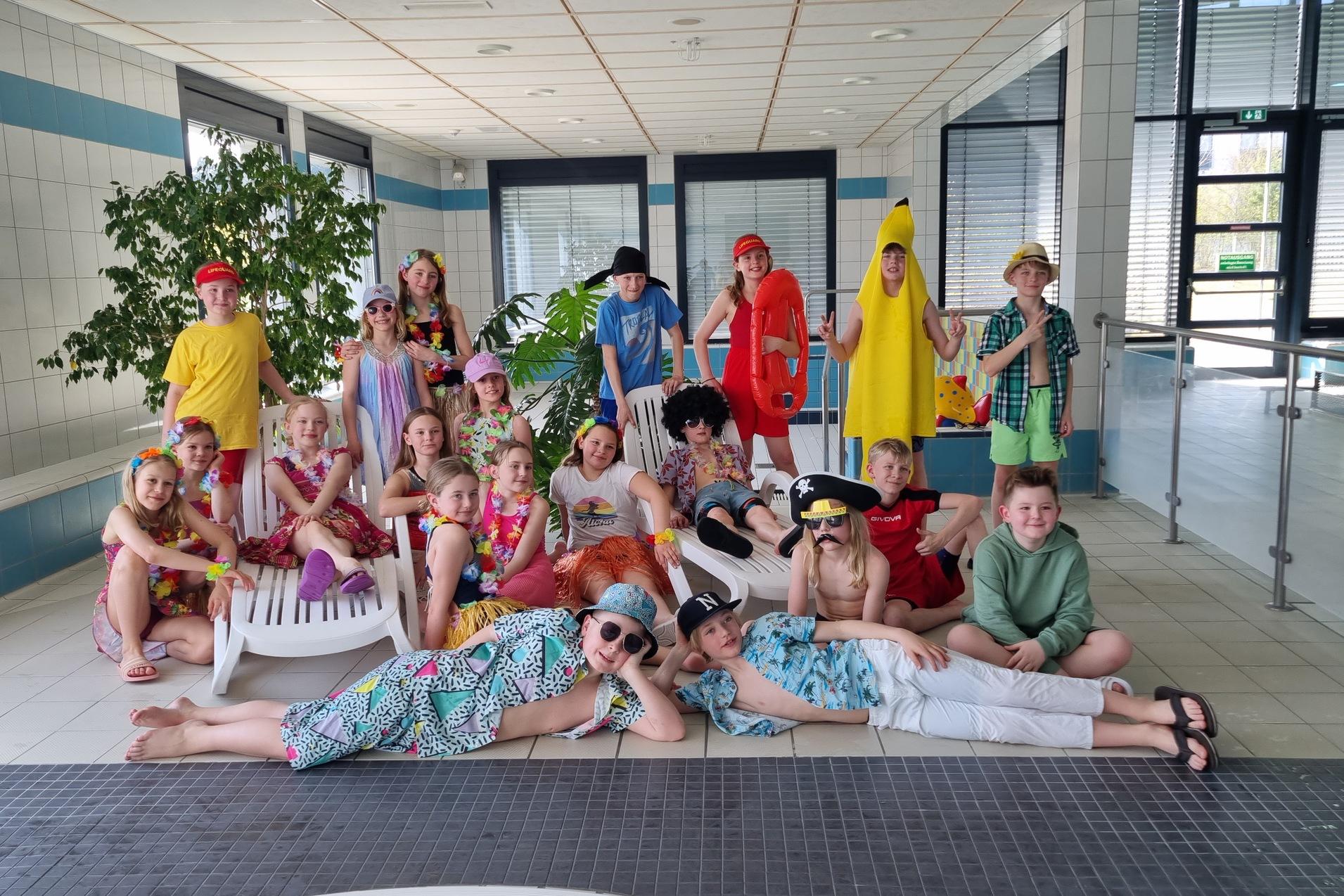 Gruppenfoto von Kindern in bunten Kostümen und Sonnenaccessoires, posierend im Schwimmbad mit Pflanzen im Hintergrund.
