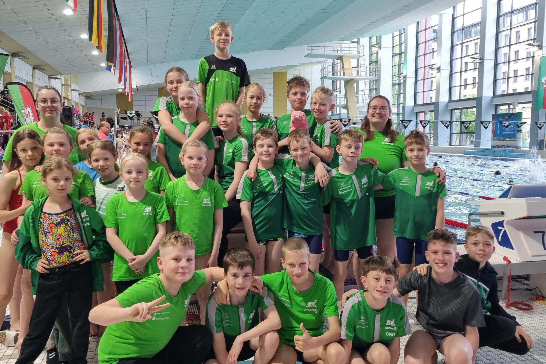 Gruppe junger Schwimmer in grünen T-Shirts posiert fröhlich am Beckenrand eines Schwimmbades.