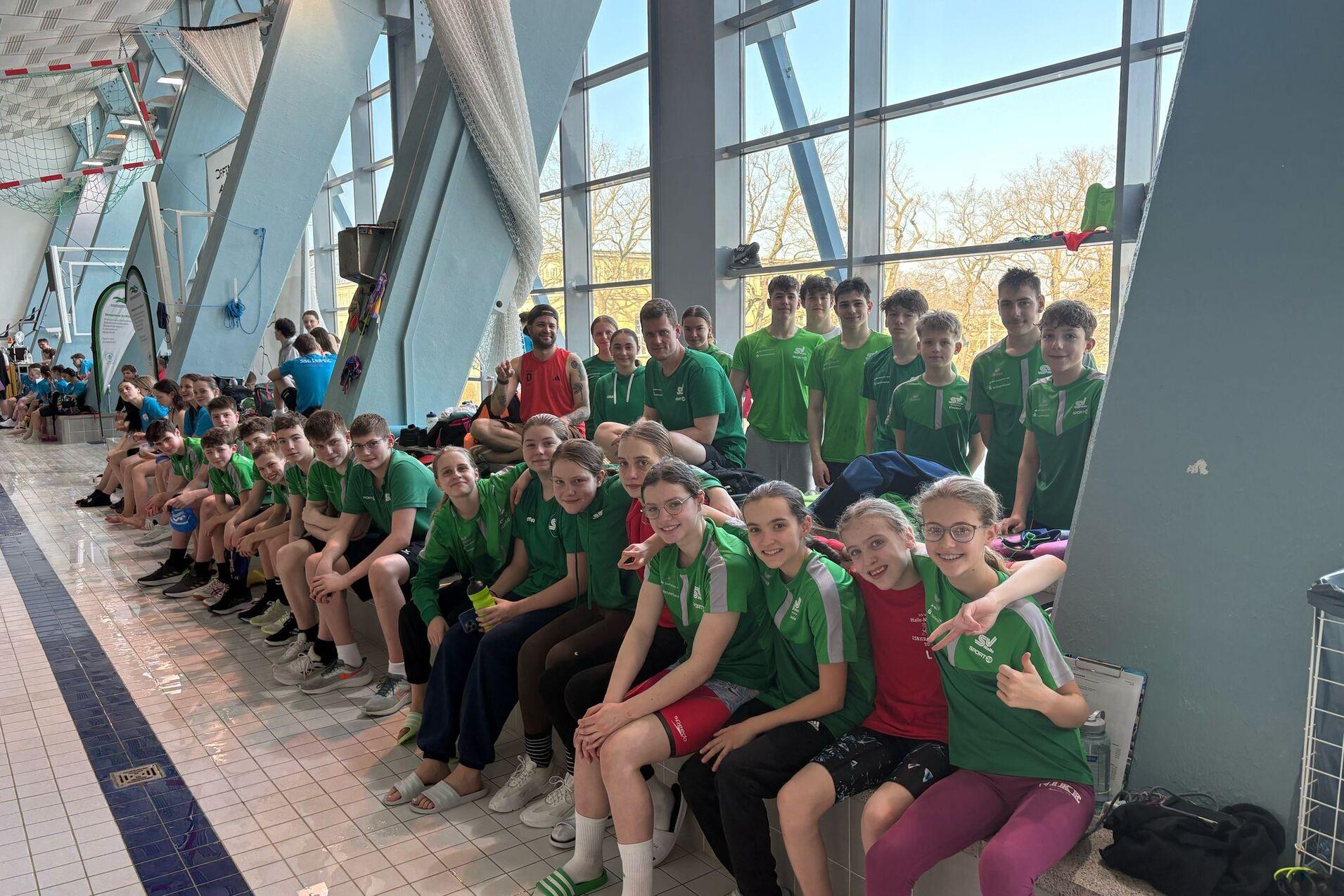 Gruppe von Schwimmern in grünen T-Shirts sitzt am Beckenrand eines Schwimmbads, einige halten Drinks und lächeln.