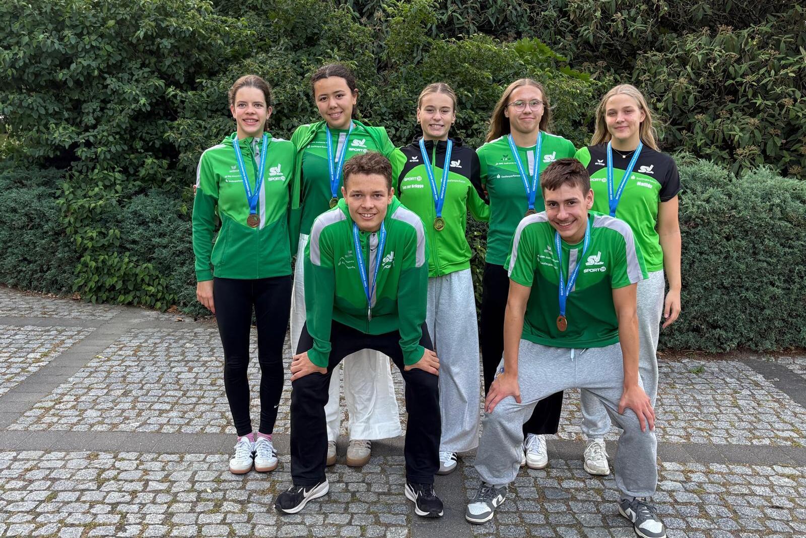 Gruppenfoto von sechs Sportlern in grünen Trikots mit Medaillen, posierend vor einer Hecke auf einem Pflasterweg.