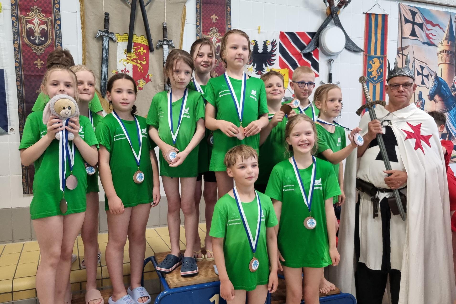 Siegerehrung Ritterschwimmen 2026 Kinder des SV Halle stehen auf dem Siegerpodest mit ihren Mediallen um den Hals. Daneben steht ein Ritter mit Rüstung und Schwert.