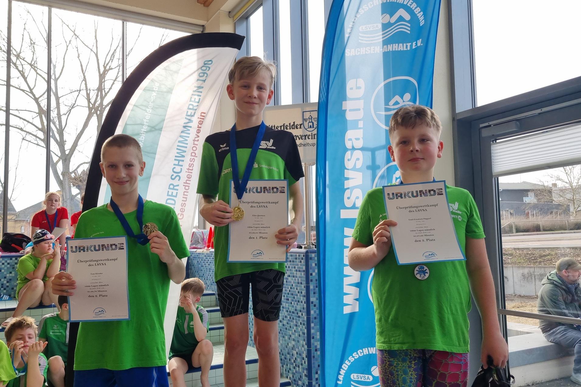 Drei Kinder in grünen Schwimmtrikots halten Urkunden und Medaillen bei einer Schwimmveranstaltung.