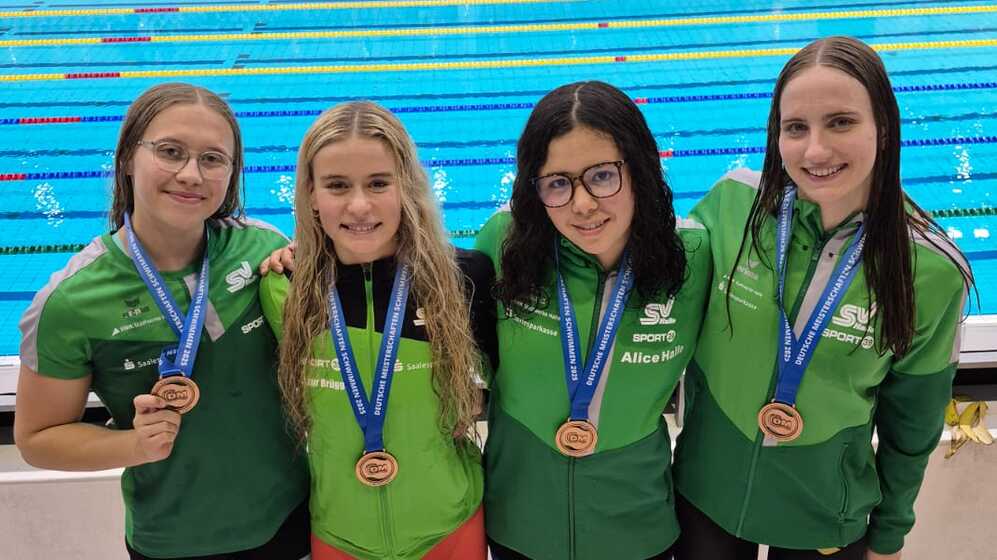 Vier Schwimmerinnen in grünen Trikots mit Medaillen posieren vor einem Wettkampfbecken.