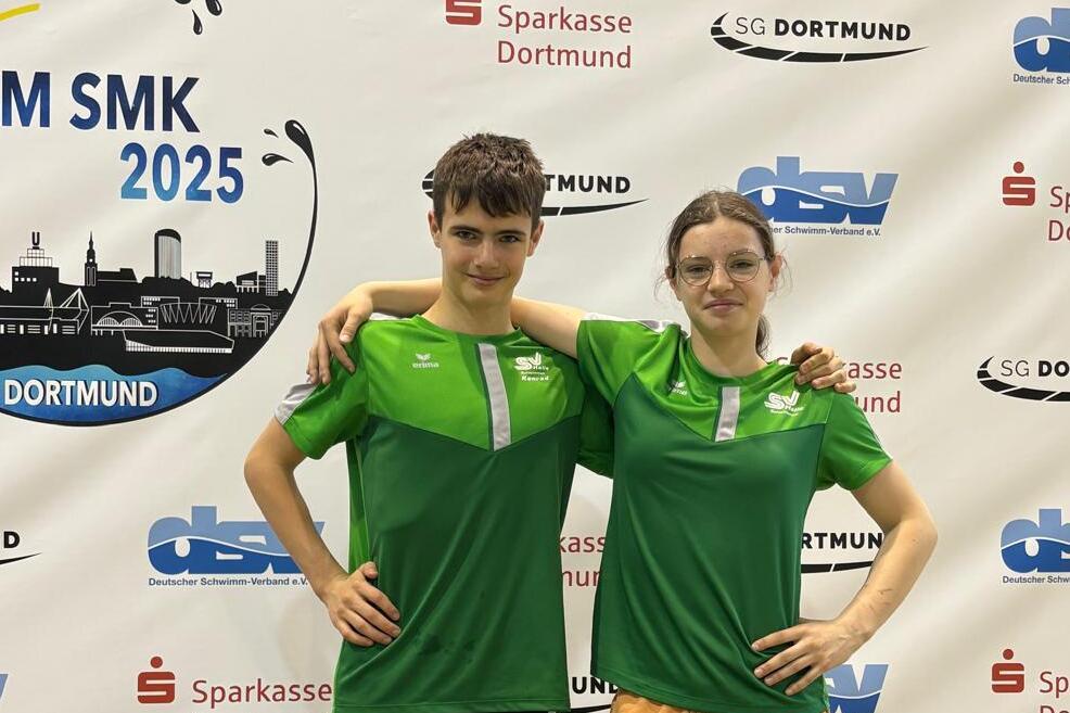 Zwei junge Schwimmer in grünen T-Shirts posieren vor einem Banner der Deutschen Meisterschaften 2025 in Dortmund.