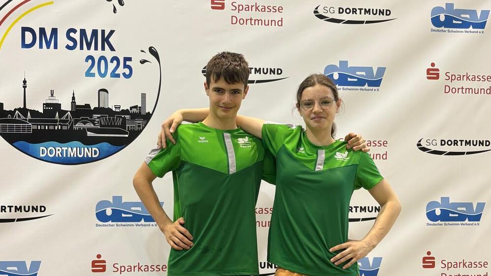 Zwei junge Schwimmer in grünen T-Shirts posieren vor einem Banner der Deutschen Meisterschaften 2025 in Dortmund.