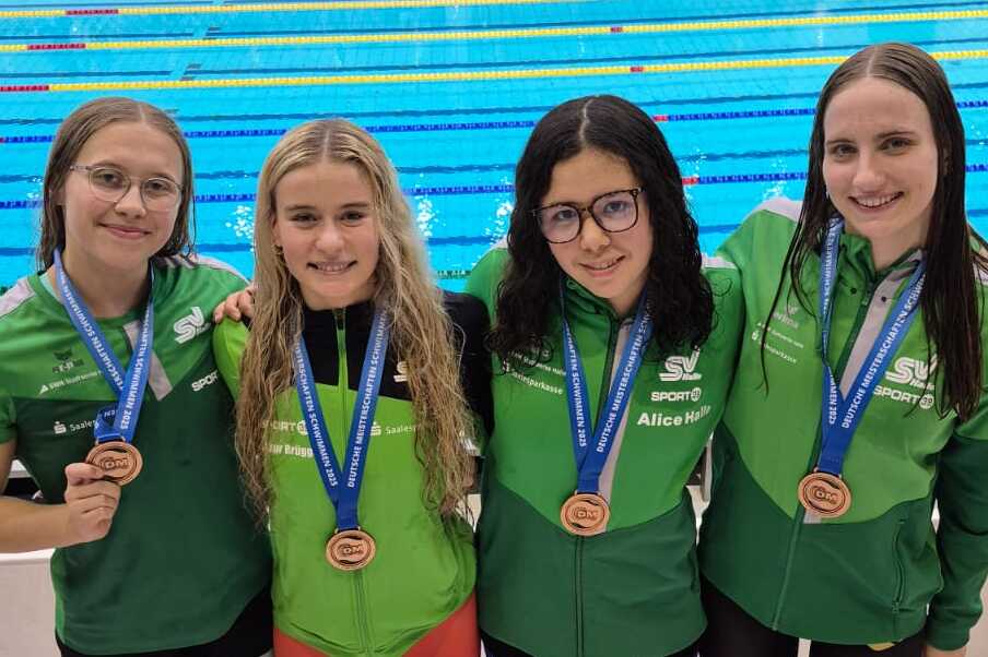 Vier Schwimmerinnen in grünen Trikots mit Medaillen posieren vor einem Wettkampfbecken.