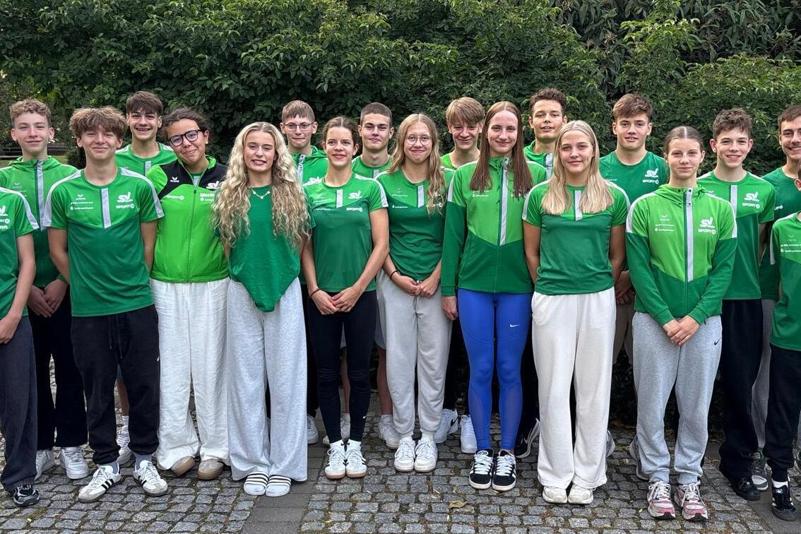 Gruppenbild von 16 jungen Sportlern in grünen Trikots, stehend vor einer grünen Hecke auf einem gepflasterten Weg.