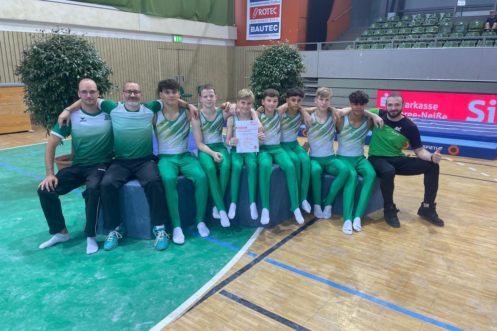 Gruppenfoto von Gymnastikern in grünen Anzügen, umgeben von Trainern, mit Urkunde in der Mitte.