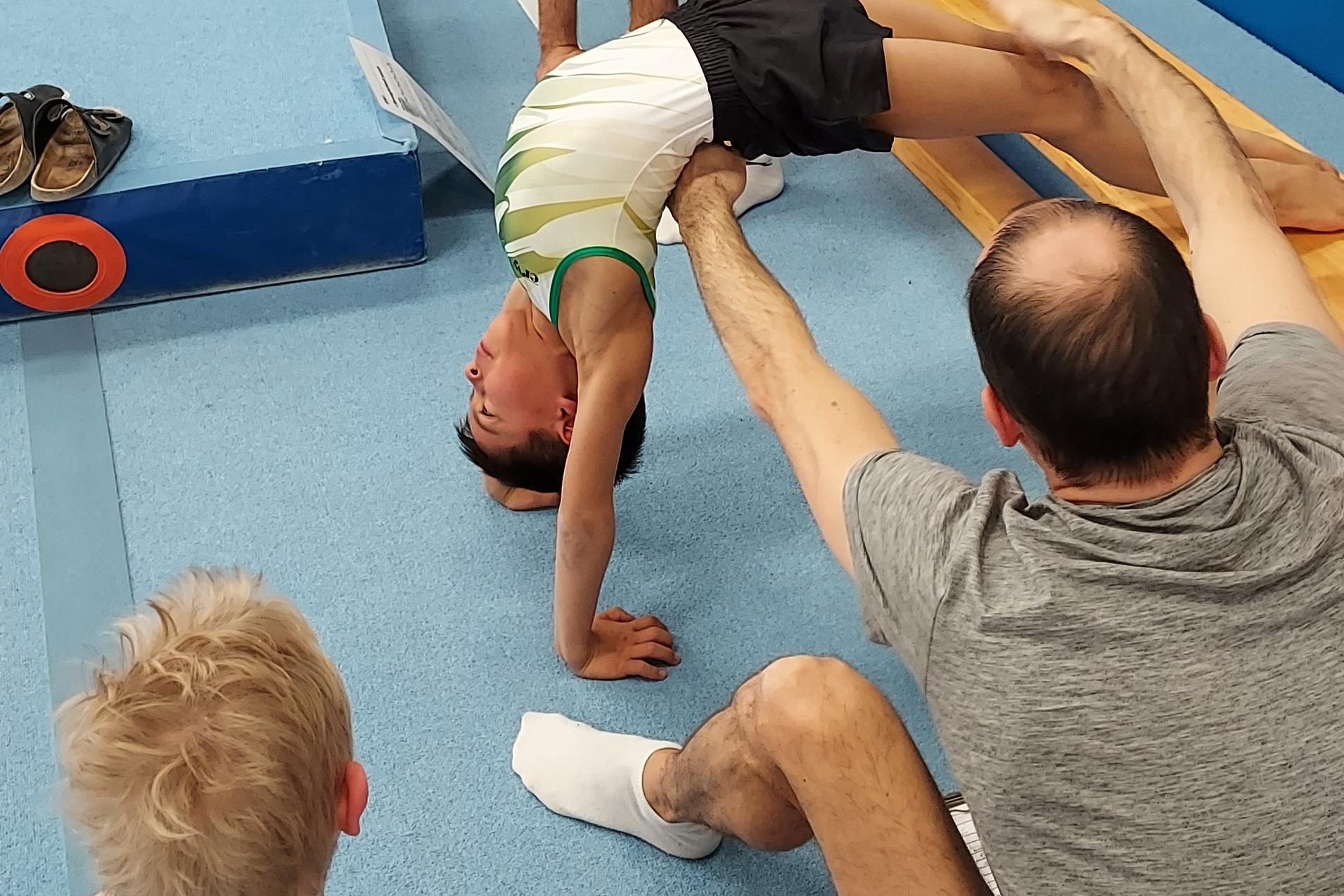 Ein Junge macht eine Brücke, während Trainer und Zuschauer im Gymnastikraum zuschauen. Matte und Turngeräte im Hintergrund.