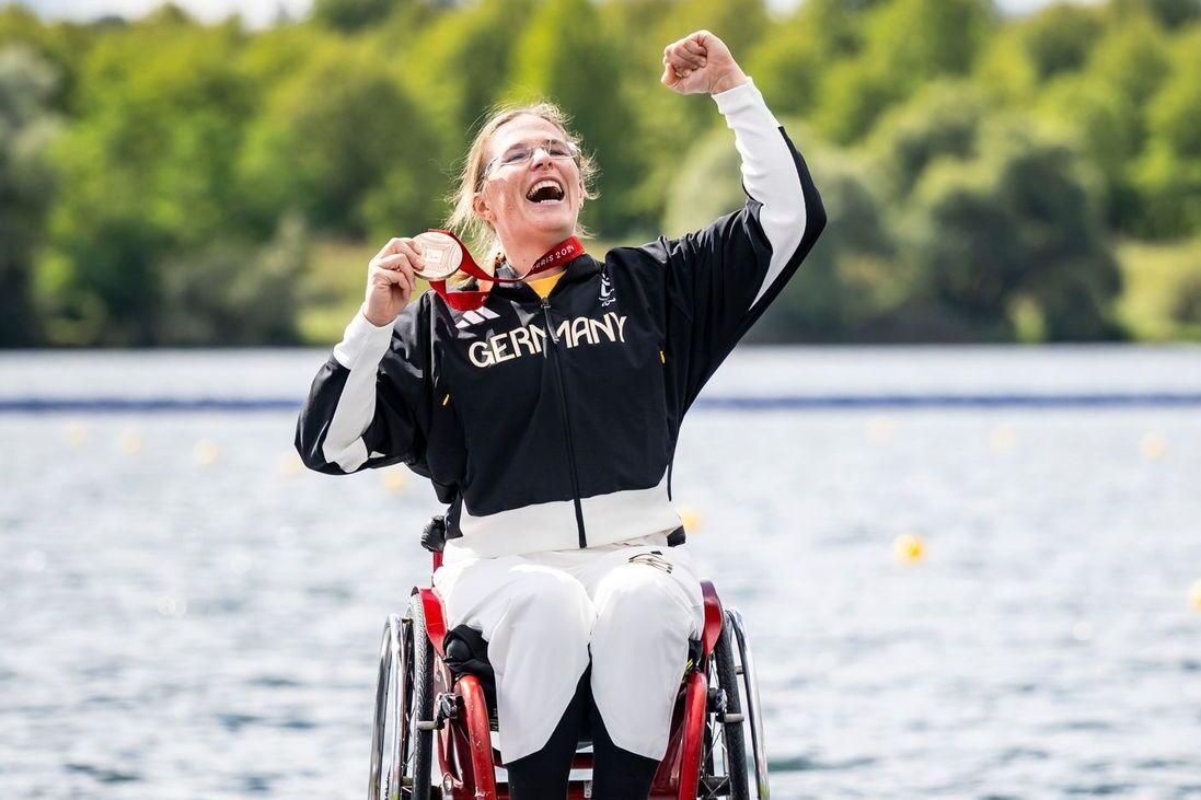Anja Adler Bronzemedaille Paris 2024 Eine Frau im Rollstuhl jubelt auf dem Siegerpodest und hält eine Medaille, während ein Mann im Hintergrund steht.