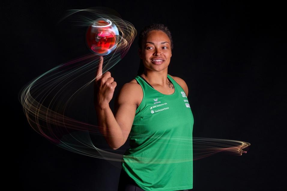 Sportlerin in grünem Trikot balanciert einen roten Ball auf dem Zeigefinger, mit dynamischen Lichtspuren im Hintergrund.