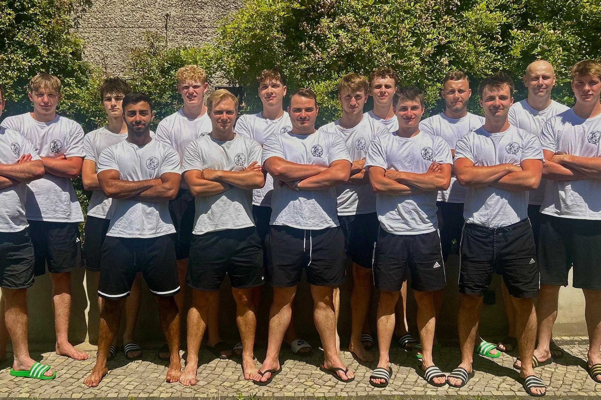 Gruppe von zwölf jungen Männern in weißen T-Shirts und schwarzen Shorts, posed vor einer grünen Hecke im Freien.
