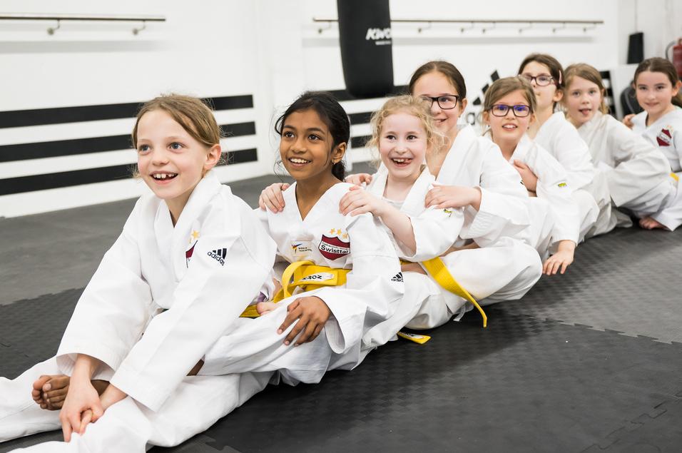 Gruppe von lächelnden Mädchen in weißen Karate-Anzügen sitzt auf einer Matte in einem Trainingsraum.